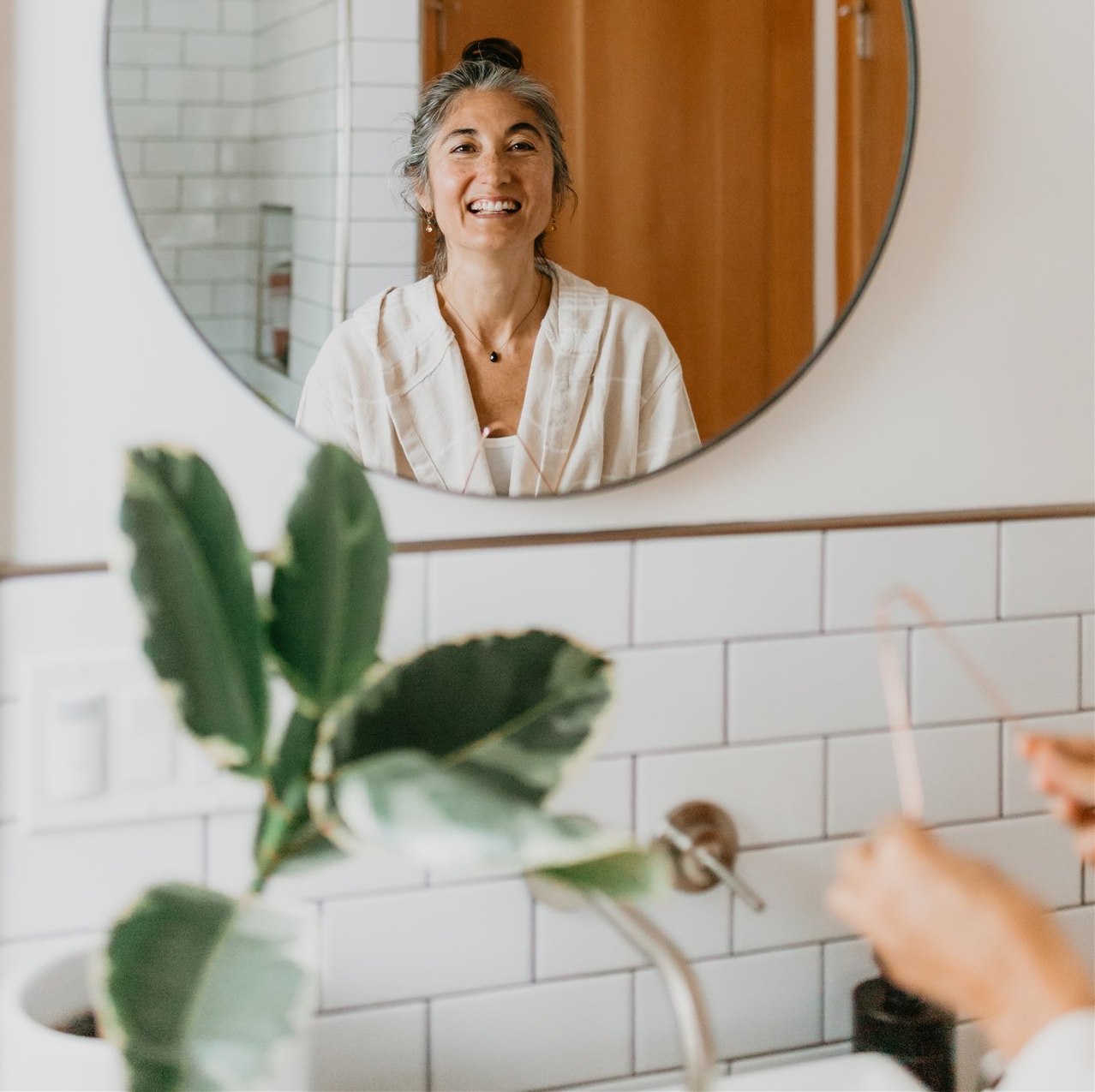 Smiling woman reflected in bathroom mirror representing Ayurvedic consultations online by DC-PAAVANI Ayurveda