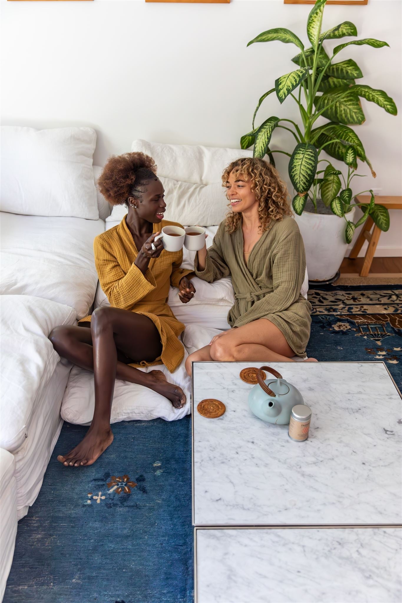 Two women sitting on couch enjoying organic loose leaf herbal tea in white mugs at home