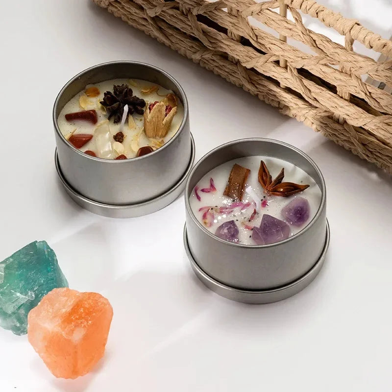 Two scented candles with dried flowers in silver tins placed on white surface next to colorful crystals