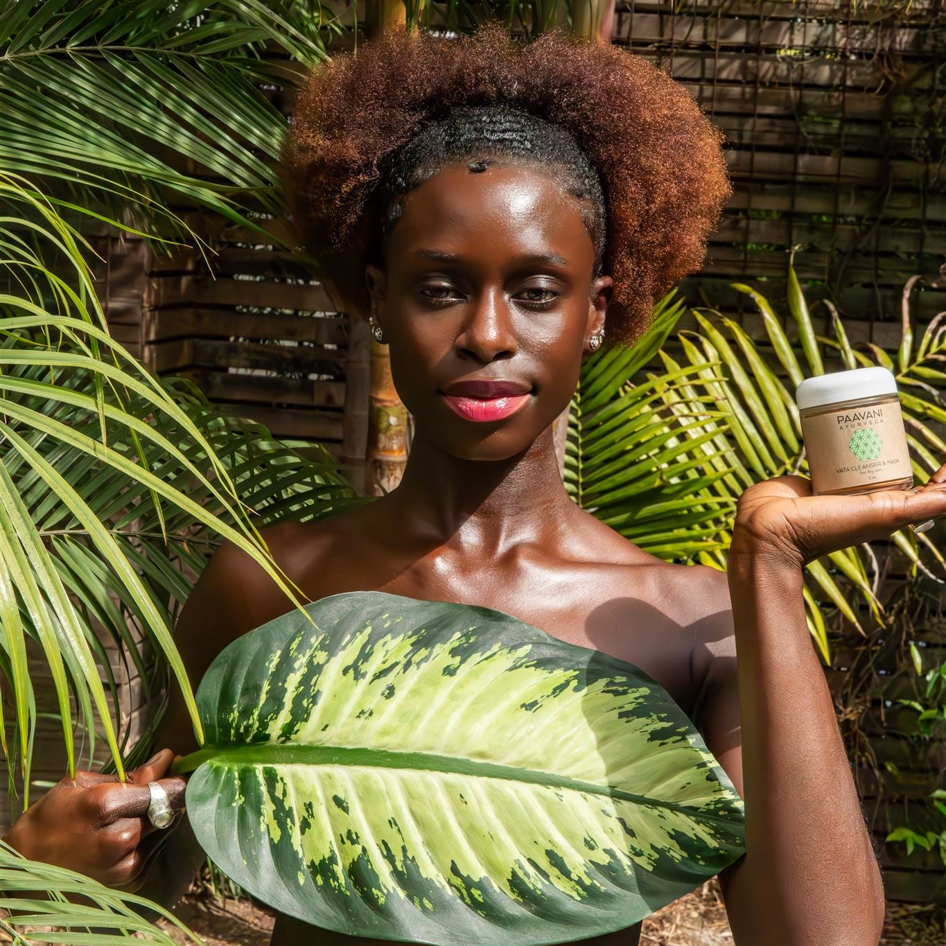 Woman holding jar of DC-PAAVANI Ayurveda ayurvedic cleanser for dry skin surrounded by green tropical leaves