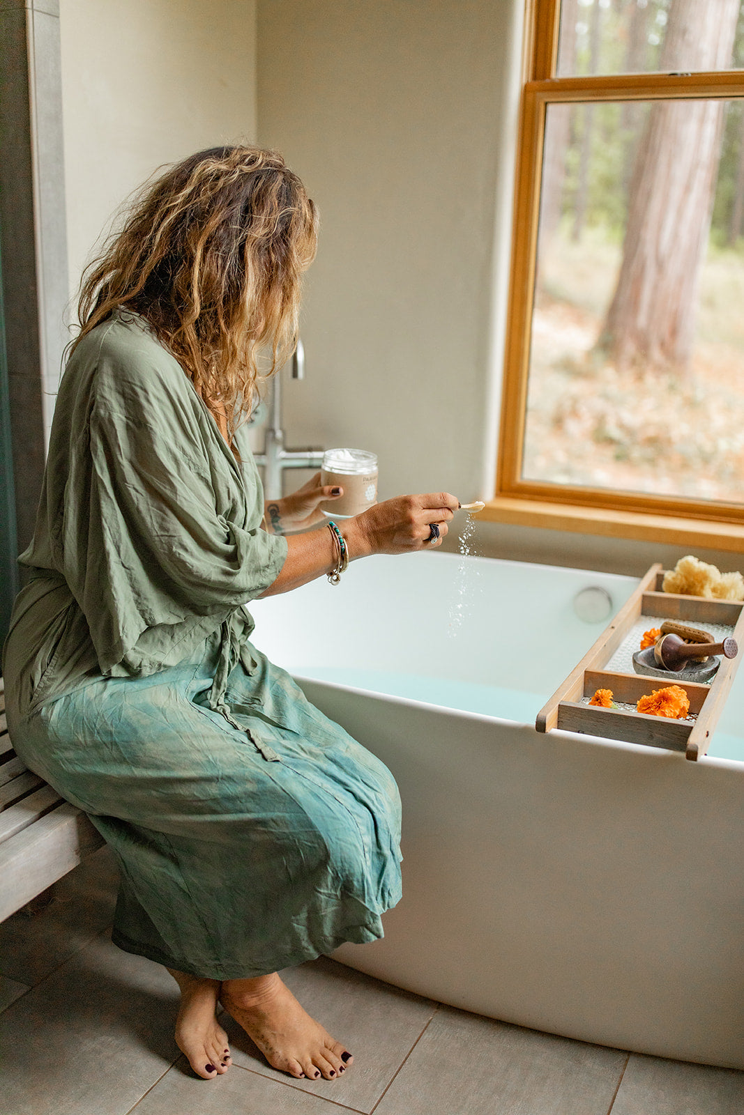 Woman in green robe preparing Ayurvedic coconut milk bath by pouring milk into white bathtub next to window