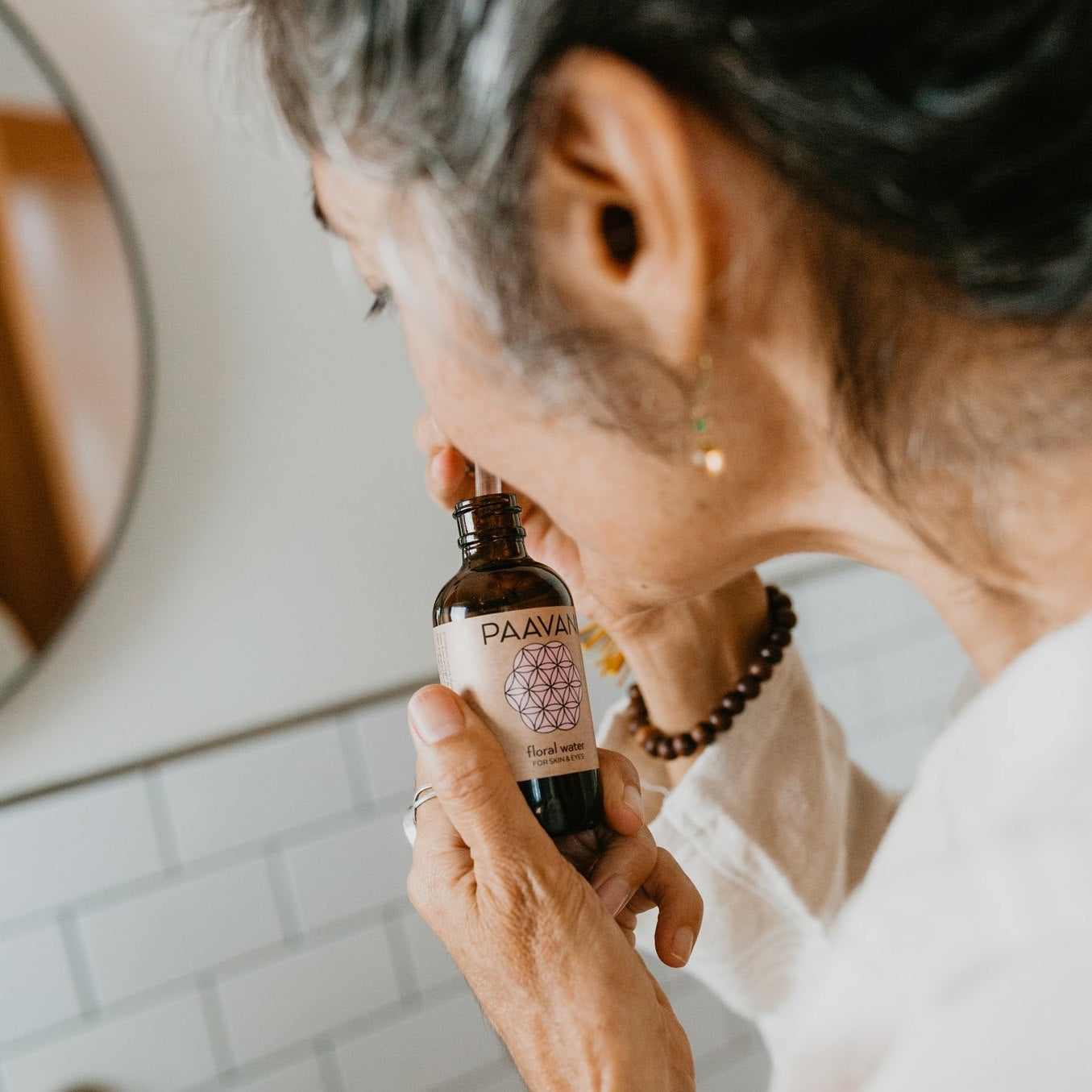 Person holding DC-PAAVANI Ayurveda bottle of botanical floral water for skin near bathroom mirror