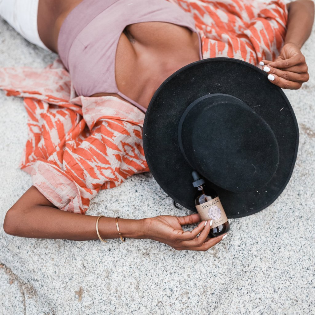 Person holding DC-PAAVANI Ayurveda botanical floral water for skin bottle while lying down with black hat and orange patterned jacket