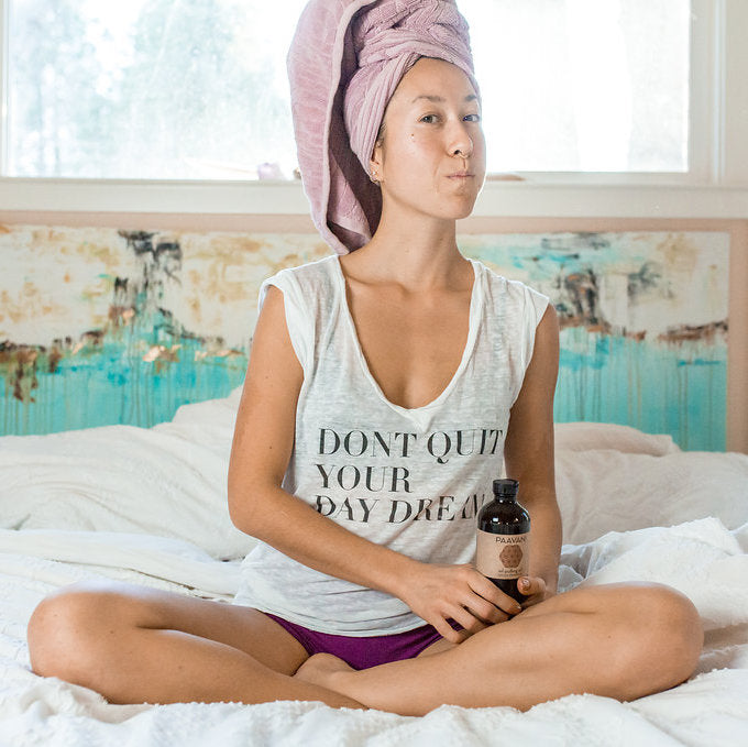 Woman sitting cross-legged on bed holding Dinacharya ritual kit for daily wellness bottle in a relaxed home setting