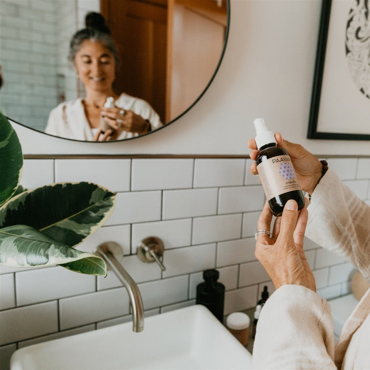 Woman holding a bottle from DC-PAAVANI Ayurveda tridoshic facial ritual kit near bathroom sink mirror